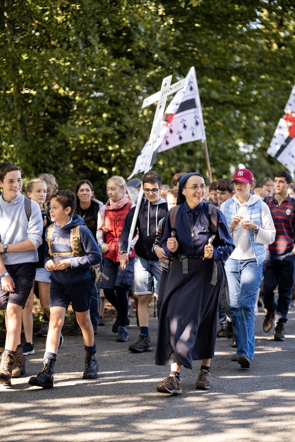 En gruppe pilegrimer vandrer under trær. Det er en ordenssøster foran, og mange unge folk med flagg og et kors lenger bak.